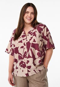 Camicia a maniche corte beige con grande motivo floreale bordeaux, dotata di colletto a punta e chiusura con bottoni, abbinata a pantaloni marrone chiaro.