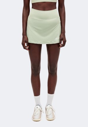 Donna che indossa una gonna sportiva verde chiaro e un top abbinato, calzini bianchi e scarpe da ginnastica bianche, in piedi contro uno sfondo semplice.