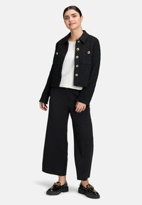 Giacca in tweed nera con bottoni dorati, dotata di colletto e due tasche frontali, abbinata a una maglietta bianca e pantaloni neri a gamba larga. Scarpe nere.