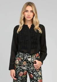 Blouse noire avec des volants sur le devant, manches longues et fermeture à boutons. Associée à un pantalon multicolore à motifs floraux.