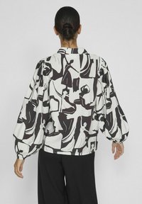 Blusa de manga larga en tela blanca con un patrón abstracto negro. Presenta mangas abullonadas y un cuello con solapa. Detalles adicionales incluyen puños elásticos.