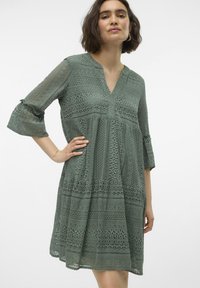 Robe en dentelle verte avec un design à volants, décolleté en V et manches 3/4. Présente des motifs complexes et un tissu texturé pour un détail supplémentaire.