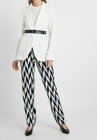 Valkoinen bleiseri mustalla vyöllä, yhdistettynä kuviollisiin housuihin, joissa on laivastonsinisiä ja mintunvihreitä geometrisia muotoja, sekä mustat kengät.