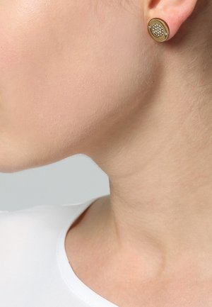 Gros plan sur le cou et la ligne de la mâchoire d'une personne portant un haut blanc et une boucle d'oreille clou en or incrustée de petits cristaux clairs.