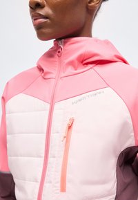 Gros plan d'une personne portant une veste à capuche rose, blanche et marron avec une fermeture éclair frontale et une fermeture éclair sur la poche poitrine.
