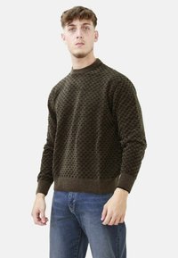 Maglione marrone a motivi con collo alto, polsini e orlo a coste. Realizzato in tessuto testurizzato con un design squadrato. Abbinato a jeans blu.