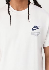 Balta medvilninė T-marškinė su tamsiai mėlynu „Nike“ logotipu ir pilkai užrašu "TIK ĮGALIOTI PERSONELIAI". Apvaliu kaklu ir trumpomis rankovėmis.