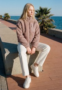 Ung kvinde iført beige fleecejakke og hvide bukser sidder på en betonmurstensrand ved havet, med palmer og klar himmel i baggrunden.