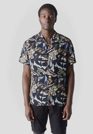 Joven de pie, vistiendo una camisa de botones de manga corta con patrones florales abstractos y pantalones cortos negros, sobre un fondo liso.