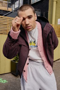 Giacca trapuntata prugna con colletto rosa, felpa grigia con grafica di un limone e pantaloni della tuta grigio chiaro. Gli accessori includono collane a strati.