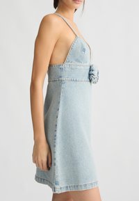 Abito in denim azzurro chiaro con corpino aderente, scollatura a V profonda, spalline sottili e dettaglio floreale in vita. Tessuto liscio e strutturato.