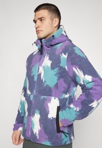 Fleece bunda v mnohých odtieňoch fialovej, modrej a bielej. Obsahuje zips vpredu, vysoký golier a bočné vrecká s čiernymi akcentmi.