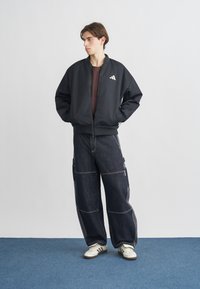 Schwarze Bomberjacke mit Frontreißverschluss und weißem Logo, kombiniert mit dunklen Denim-Cargohosen mit seitlichen Reißverschlüssen und weißen Nähten. Weiße Sneaker.