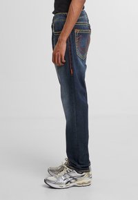 Jeans de un azul oscuro con costuras amarillas, bolsillos laterales y un diseño único en el bolsillo trasero. Combinados con zapatillas deportivas grises y plateadas.