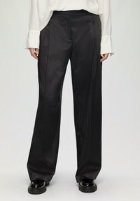 Schwarze, weit geschnittene Hosen aus einem glatten, satinähnlichen Material. Mit hohem Bund und scharfen vorderen Falten. Kombiniert mit klobigen schwarzen Schuhen.