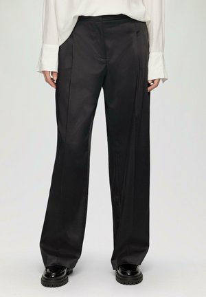 Schwarze, weit geschnittene Hosen aus einem glatten, satinähnlichen Material. Mit hohem Bund und scharfen vorderen Falten. Kombiniert mit klobigen schwarzen Schuhen.