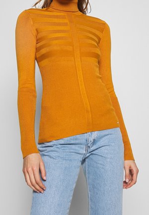 Femme portant un pull à col roulé côtelé jaune moutarde avec des détails à rayures horizontales, associé à un jean bleu clair, sur un fond uni.