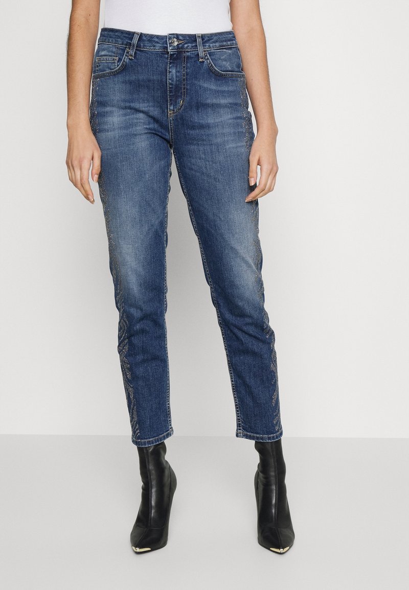 Jeans de mezclilla en azul oscuro con corte ajustado, que presentan adornos detallados a lo largo de las costuras laterales y un diseño clásico de cinco bolsillos.