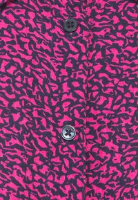 Primo piano di un tessuto rosa brillante con un motivo astratto nero irregolare e due bottoni neri a quattro fori allineati verticalmente.