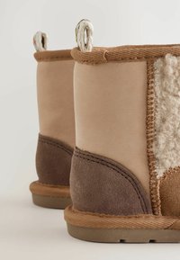 Ein Paar beige und braune Winterstiefel mit Zuglaschen, strukturiertem Stich und weichen, wollähnlichen Seitenpaneelen auf einem neutralen Hintergrund.