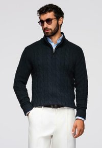 Homme portant un pull en maille torsadée à col zippé sombre sur une chemise bleu clair et un pantalon blanc, avec des lunettes de soleil, posant devant un fond gris.