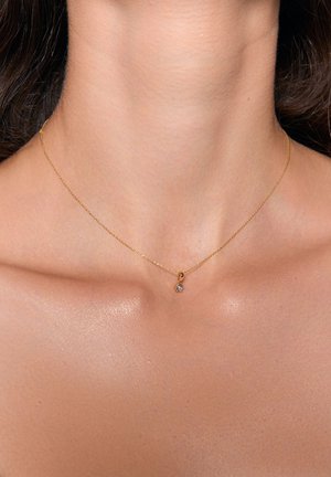 Złoty naszyjnik z delikatnym łańcuszkiem i małym wisiorkiem z przezroczystym kamieniem szlachetnym, spoczywający na gładkim, nagim dekolcie.