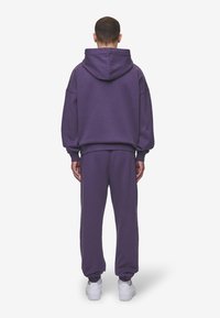 Felpa viola con cappuccio, vestibilità oversize, polsini a coste e pantaloni felpa viola abbinati. Sneakers bianche completano l'outfit. Vista posteriore.