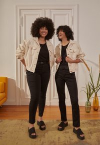 Duas pessoas vestem camisetas pretas, calças pretas e jaquetas de denim bege. Elas têm cabelo encaracolado e usam sandálias pretas de plataforma. Um sofá amarelo e plantas são visíveis.