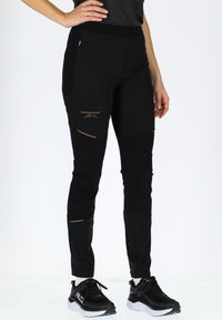 Black softshell pants with gold accents and logo, featuring zippered pockets and a fitted design for ease of movement.