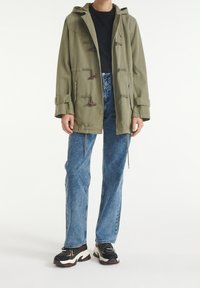 Khaki park z kapturem, zapinany na zapięcia typu toggle, z długimi rękawami i kieszeniami bocznymi. Noszony na czarnej koszulce i jasnoniebieskich dżinsach prostych.