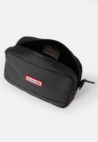 Čierna obdĺžniková taška vyrobená z odolného materiálu. Obsahuje zips, vnútorné podšívku a viditeľné červené a biele logo na prednej strane.