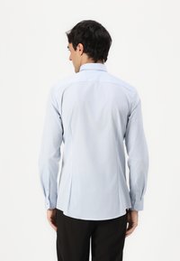 Cămașă albastru deschis cu mânecă lungă, cu tiv curbat, design cu pliuri pe spate și manșete cu nasturi. Textură de țesătură netedă, potrivire ajustată.