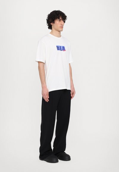 T-shirt blanc à manches courtes avec un design graphique bleu et rouge sur la poitrine, associé à un pantalon noir ample et des chaussures noires.