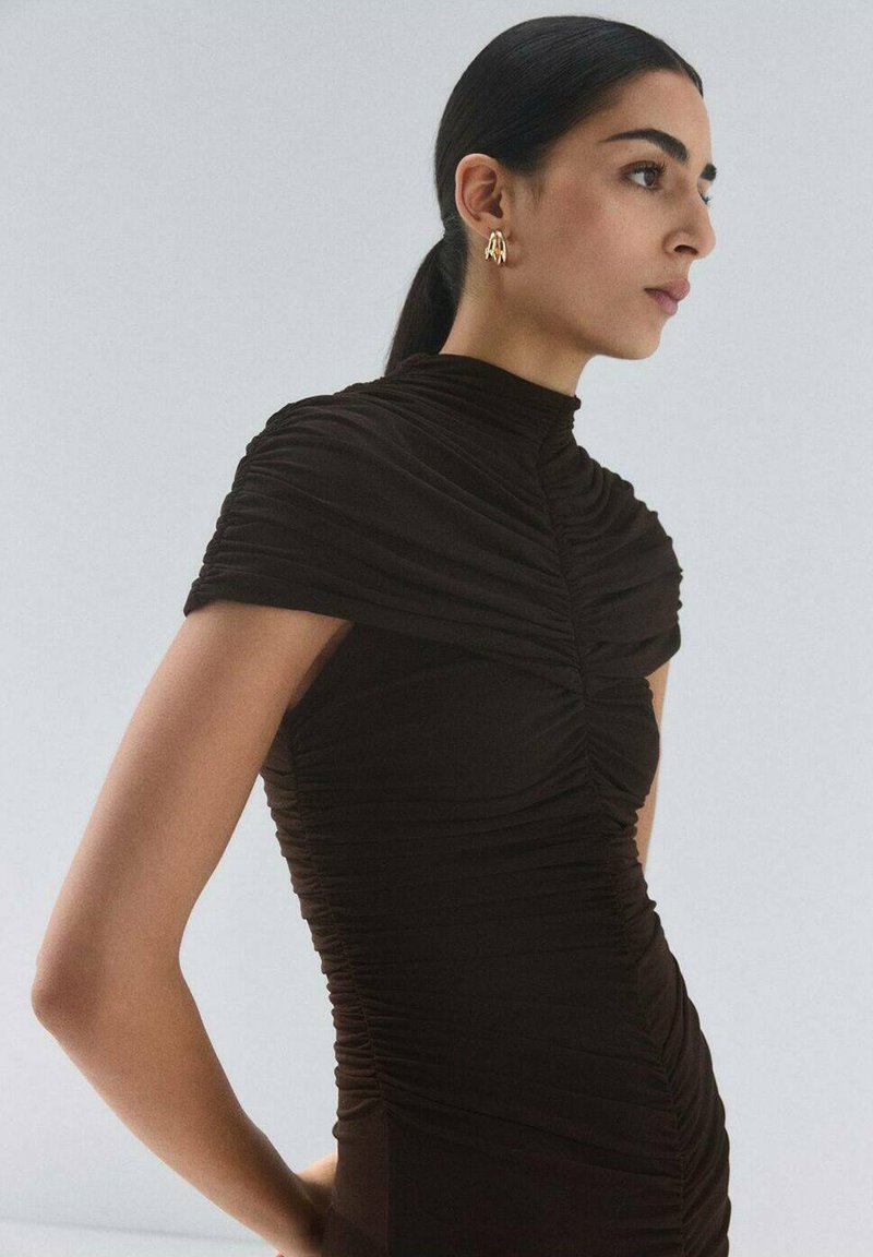 Robe noire froncée avec un col montant et des manches courtes, présentant une texture lisse et une coupe près du corps, complétée par des boucles d'oreilles dorées.