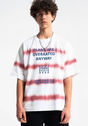 Młoda osoba z kręconymi włosami nosi białą i czerwoną koszulkę w paski z niebieskim napisem: „PLANY I TAK SĄ PRZEREKLAMOWANE” oraz „ZOSTAW MIEJSCE NA NIEOCZEKIWANE.”