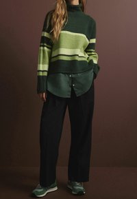 Gestrickter Pullover mit grünen und hellgrünen Streifen und einem schwarzen Rollkragen; darüber liegt ein dunkelgrünes, langärmliges Hemd mit Knöpfen und schwarze Hose.