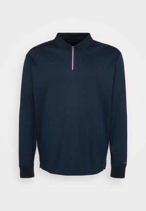 Tamsiai mėlyna polo marškinė su ilgomis rankovėmis, užtrauktuku ir raudonos, baltos bei mėlynos spalvų juostelėmis ant užtrauktuko traukimo, bei mažas logotipas ant rankogalių.