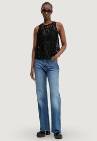 Top senza maniche in pizzo nero con motivi floreali, abbinato a jeans a zampa d'elefante blu chiaro. La modella sta in piedi con sicurezza, con le mani nelle tasche. Scarpe nere.