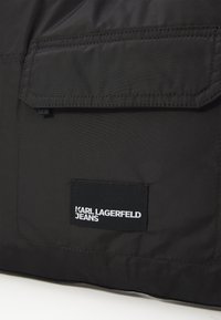 Juodas audinio krepšys su atlenkiama kišenė. Audinė etiketė rodo "KARL LAGERFELD JEANS" baltomis raidėmis juodame fone. Lygus paviršius.