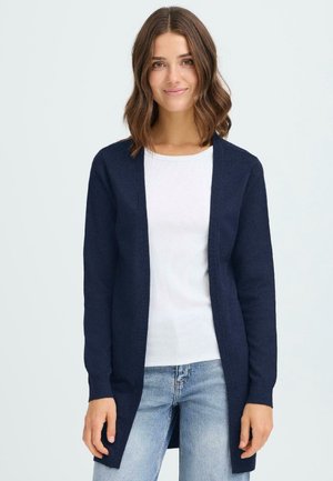 Mörkblå lång kofta i mjukt stickat material, öppen frontdesign, ribbade ärmslut, bärs över en vit skjorta och ljusblå jeans.