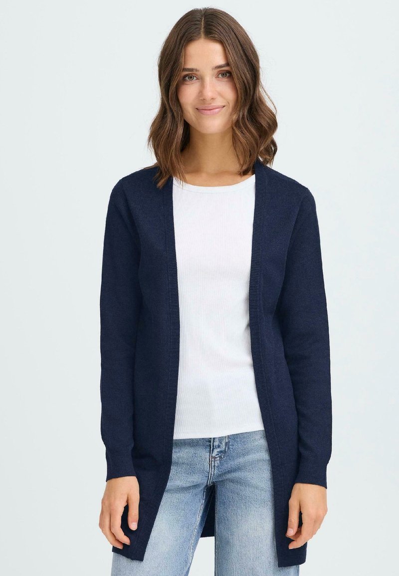 Cardigan long bleu marine en tricot doux, design ouvert à l'avant, poignets côtelés, porté sur une chemise blanche et un jean bleu clair.