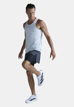 Atletisk mand iført hvidt tanktop, sorte shorts og hvide sneakers løfter det ene ben, mens han kigger ned mod en ensfarvet baggrund.