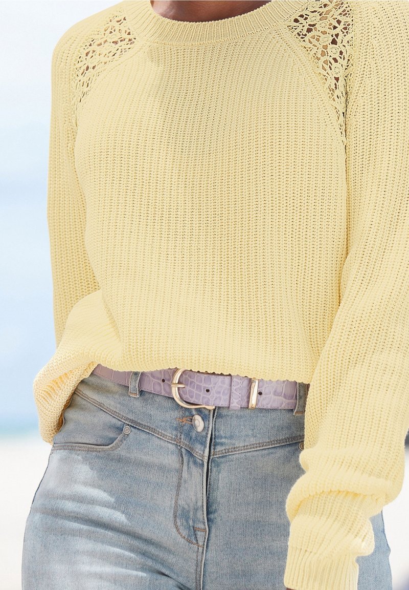 Żółty dziergany sweter z koronkowymi akcentami na ramionach, zestawiony z jasnoniebieskimi, wysokimi dżinsami i jasnofioletowym paskiem.