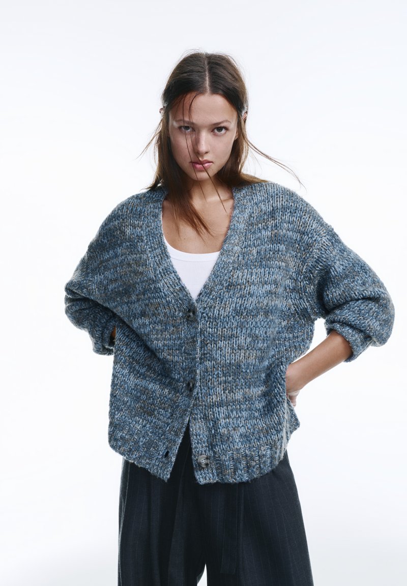 Gestrickte Strickjacke in Blau mit beigen Sprenkeln, mit tiefem V-Ausschnitt, großen Knöpfen und übergroßen Ärmeln. Strukturierter Stoff.