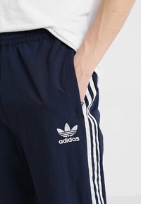 Pantalones deportivos azul marino hechos de material ligero, con rayas blancas a los lados y un logo bordado en el muslo izquierdo.