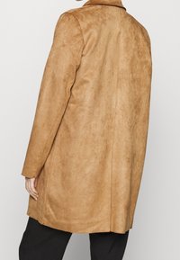 Tan mockaskinnjacka med en slät textur, långa ärmar och rak silhuett. Har en klassisk krage och når ner under höfterna.