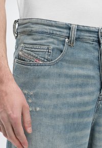 Κοντινό πλάνο ενός ατόμου που φοράει ξεθωριασμένο ανοιχτό μπλε τζιν με μικρή ετικέτα λογότυπου στην μπροστινή τσέπη και ένα άσπρο πουκάμισο που έχει μέσα στην βράκα.