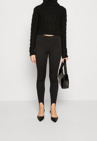 Schwarzer gestrickter Rollkragenpullover, cropped Design; schwarze Leggings mit Ausschnitten an den Knöcheln; schwarze spitze High Heels; kleine schwarze Handtasche.