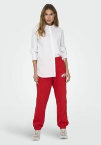 Hvid skjorte med knapper, båret over røde sweatpants med elastiske manchetter og talje. Lysegrå sneakers med tekstureret sål. Minimalistisk design.