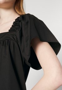 Femme portant une robe noire avec un décolleté carré froncé et de larges manches courtes fluides sur un fond clair uni.